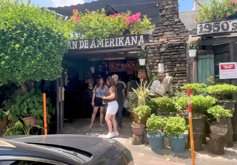 Front entrance of Pan de Amerikana restaurant in Marikina, with guests exiting under a canopy of greenery and flowers