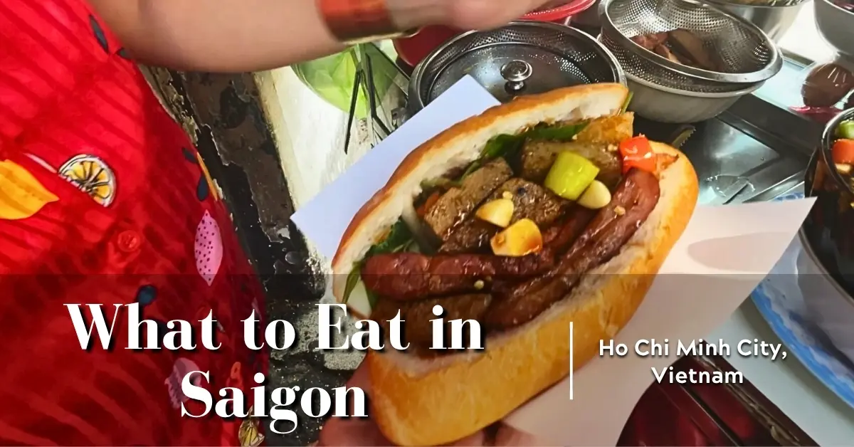 Close-up of a street vendor in Ho Chi Minh City holding a freshly made bánh mì stuffed with assorted meats, vegetables, and garlic cloves, with metal bowls of ingredients visible in the background.