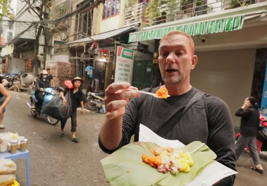 Geoff eats colourful fruit-topped sticky rice from a banana leaf plate on a Hanoi street